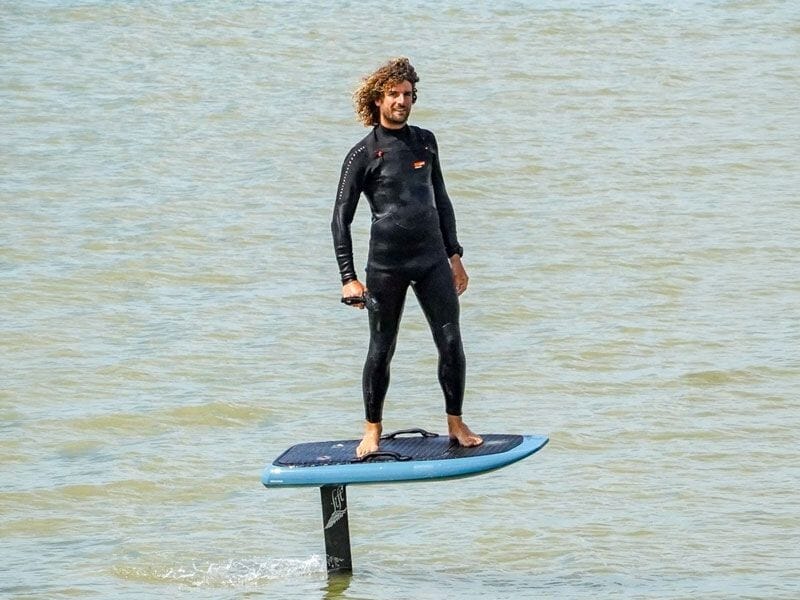 Un homme en combinaison noire debout sur une planche de surf électrique sur une eau calme, souriant.