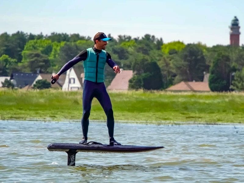 Un homme en combinaison bleue et gilet debout sur une planche de surf électrique avec un phare visible au loin.