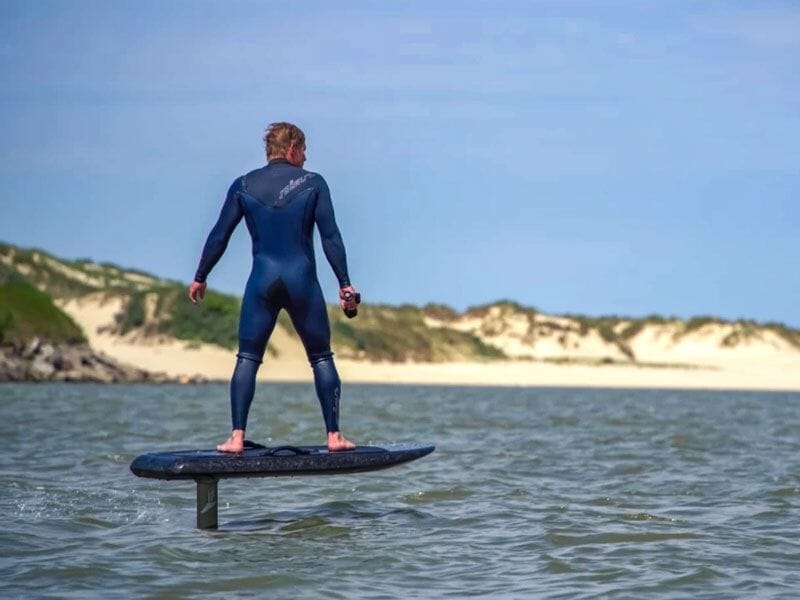 Un homme en combinaison bleue debout sur une planche de surf électrique sur une mer calme avec des dunes en arrière-plan.