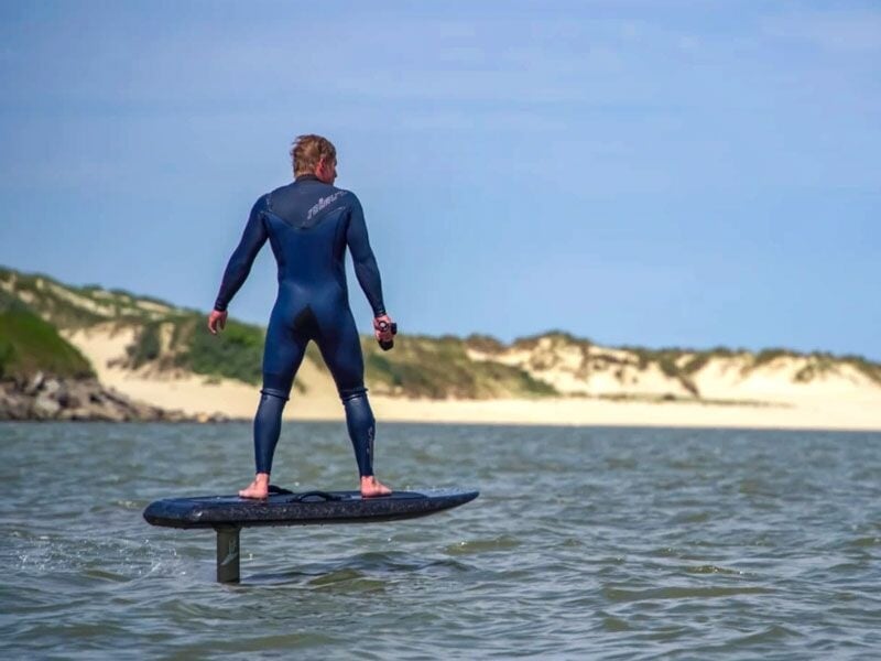 Un homme en combinaison bleue debout sur une planche de surf électrique sur une mer calme avec des dunes en arrière-plan.