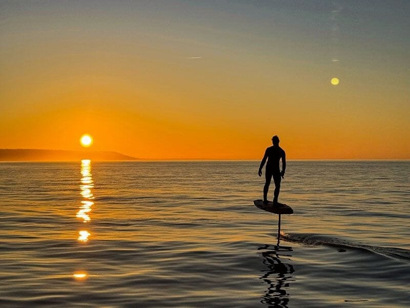 Personne sur un surf électrique glissant sur l'eau calme au coucher du soleil avec un ciel orange et reflet doré.