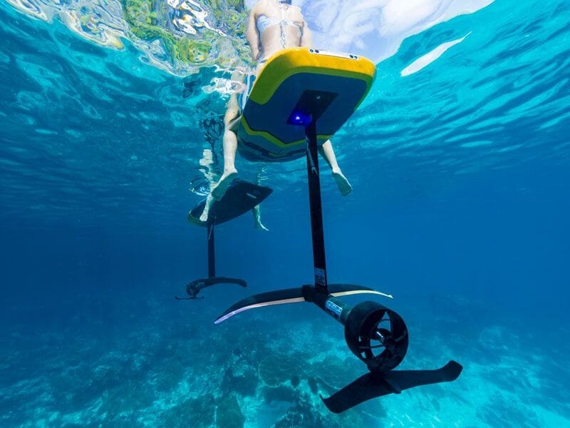 Vue sous-marine d'une personne sur un surf électrique avec une hélice, évoluant dans une eau claire et bleue.