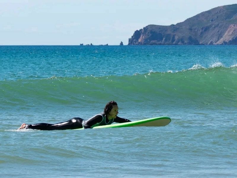Surfeur allongé sur sa planche verte, pagayant dans une vague avec une côte rocheuse en arrière-plan.