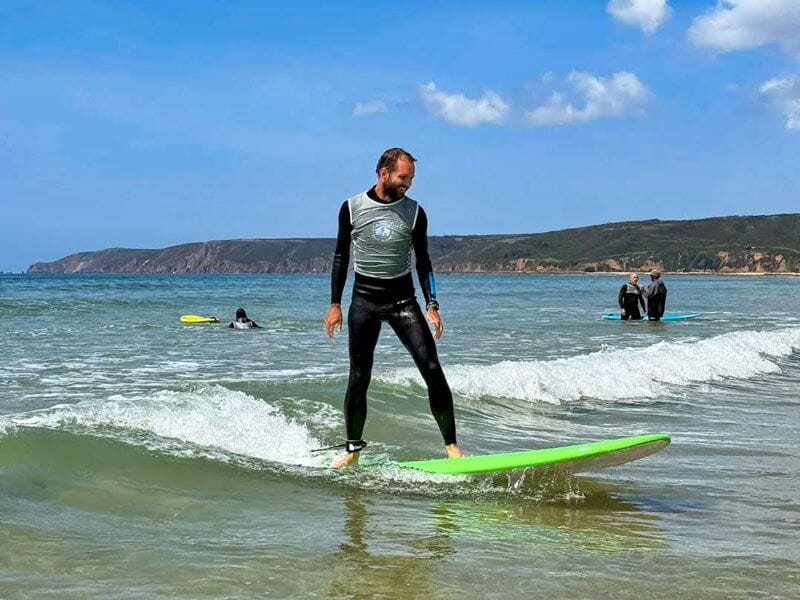 Homme en combinaison de surf debout sur une planche verte, surfant une vague calme près du rivage.