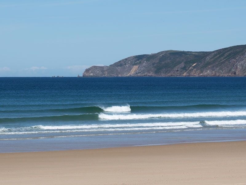 Plage de sable fin avec petites vagues déferlantes et falaises rocheuses en arrière-plan sous un ciel clair.