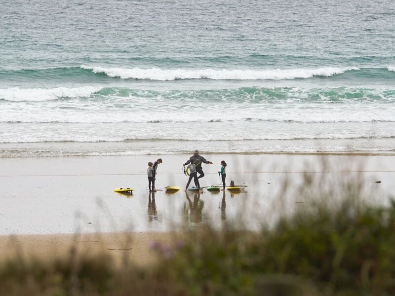 Personne en combinaison de surf avec trois enfants sur la plage, planches de surf posées sur le sable humide.