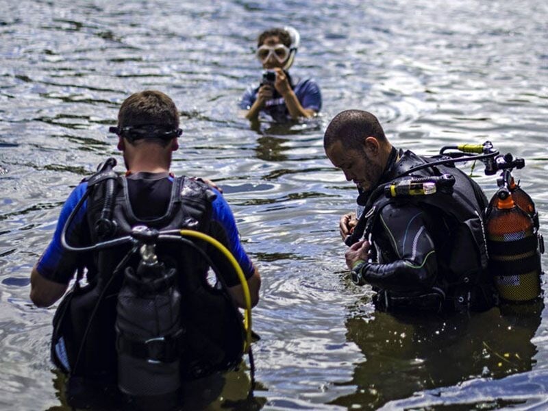 Trois plongeurs en combinaison et un enfant avec masque dans l'eau, s'apprêtant à plonger en rivière ou lac.