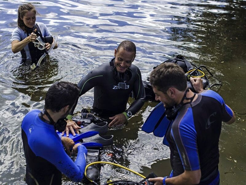 Groupe de quatre plongeurs en combinaison discutant et ajustant leur matériel de plongée dans l'eau calme.
