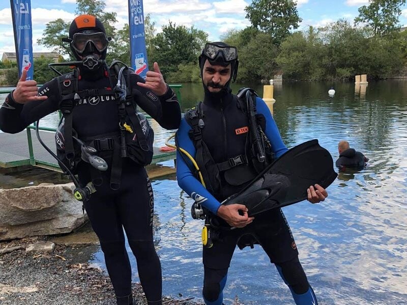 Deux hommes en combinaison de plongée posent au bord d'un lac, prêts pour un baptême de plongée sous-marine.