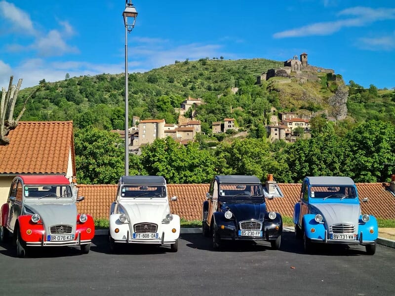 Quatre voitures Citroën 2CV colorées alignées devant un village avec une colline et un ciel bleu en arrière-plan.