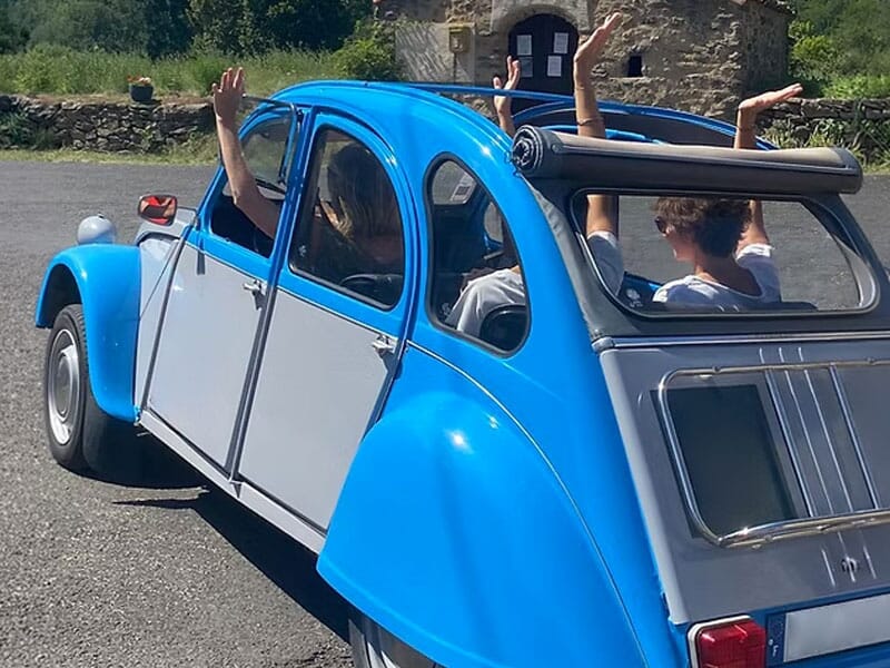 Vue arrière d'une voiture Citroën 2CV bleue avec des passagers qui lèvent les bras en signe de joie.