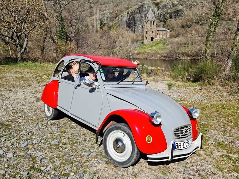 Voiture Citroën 2CV rouge et grise garée sur un chemin de cailloux avec des passagers qui saluent.