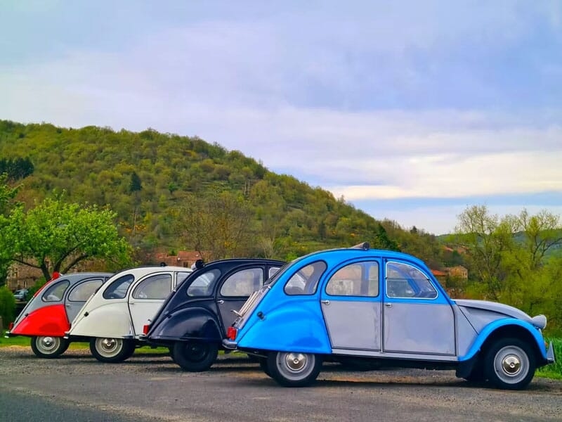 Cinq voitures Citroën 2CV alignées sur une route avec un paysage de collines et arbres verts en arrière-plan.