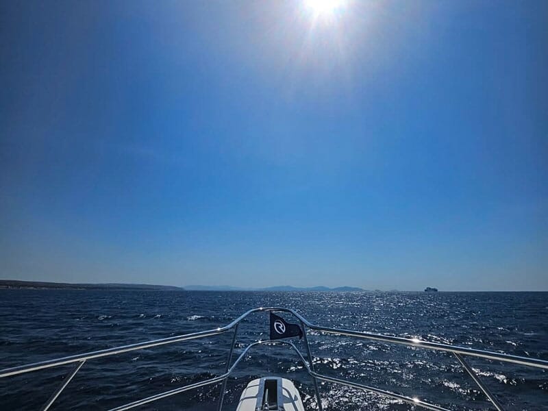 Vue depuis la proue d'un bateau sur une mer scintillante sous un ciel bleu avec un bateau au loin.