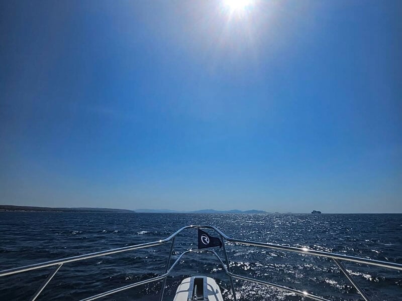 Vue depuis la proue d'un bateau sur une mer scintillante sous un ciel bleu avec un bateau au loin.