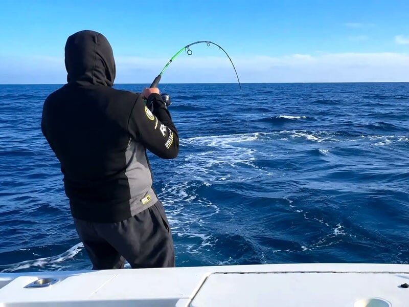 Un pêcheur en sweat à capuche noir tient une canne à pêche pliée face à la mer bleue calme.