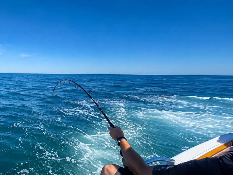 Un pêcheur assis sur un bateau tient une canne à pêche pliée avec la mer et le ciel bleu en arrière-plan.
