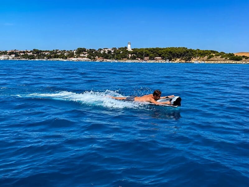 Un homme nage sur une planche dans une mer bleue avec une côte et un phare en arrière-plan sous un ciel clair.