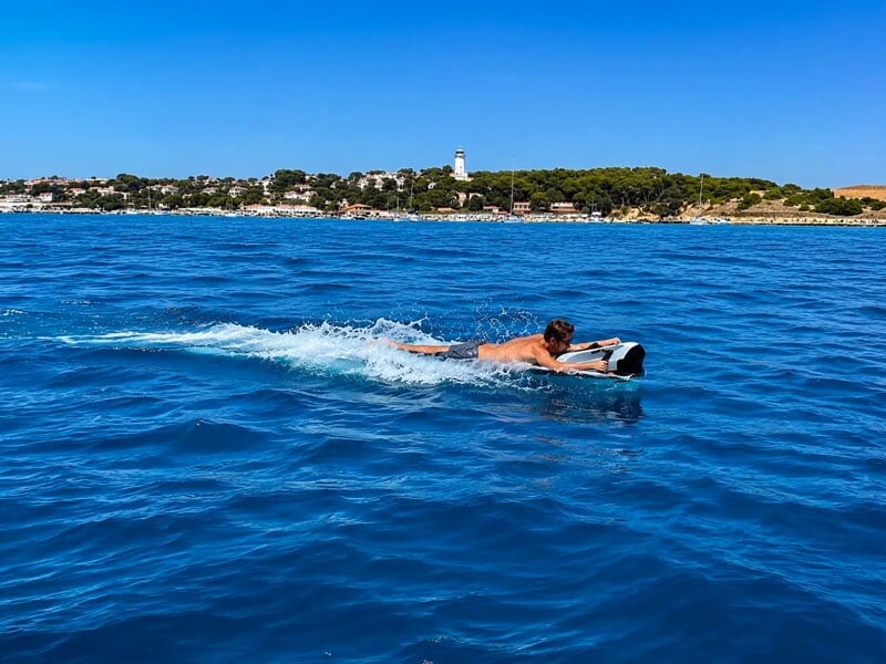 Un homme nage sur une planche dans une mer bleue avec une côte et un phare en arrière-plan sous un ciel clair.