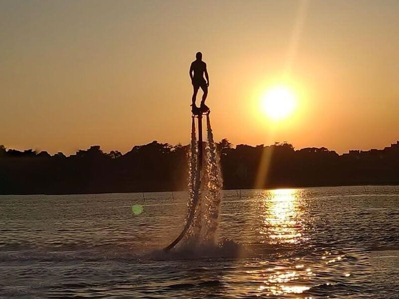 Personne en flyboard sur l'eau au coucher du soleil avec un ciel orangé et reflets lumineux sur la mer.