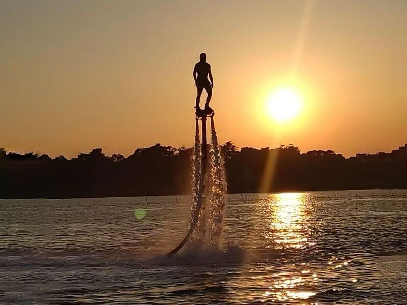 Personne en flyboard sur l'eau au coucher du soleil avec un ciel orangé et reflets lumineux sur la mer.