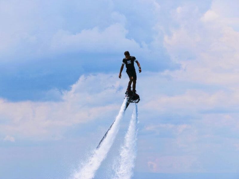Homme en flyboard s'élevant dans le ciel bleu avec des nuages blancs, propulsé par un jet d'eau puissant.