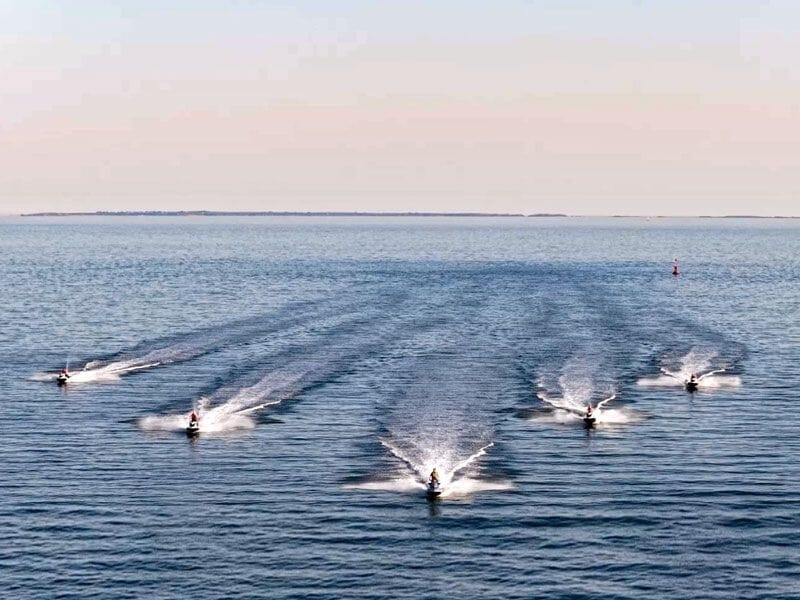Groupe de six jetskis naviguant en formation sur une vaste étendue d'eau sous un ciel clair au coucher du soleil.