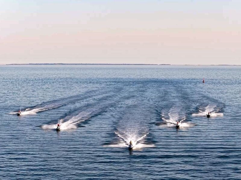 Groupe de six jetskis naviguant en formation sur une vaste étendue d'eau sous un ciel clair au coucher du soleil.