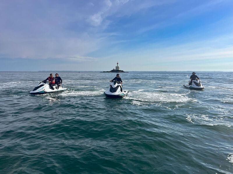 Trois personnes sur des jetskis se dirigeant vers un phare visible au loin sur une mer calme sous un ciel bleu.