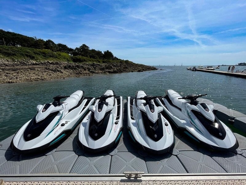 Quatre jetskis alignés sur un ponton flottant, prêts à être utilisés, avec un paysage côtier en arrière-plan.