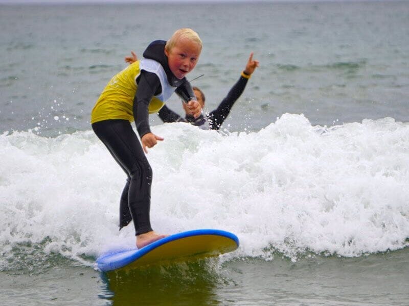Un enfant en combinaison de surf jaune et noire sur une planche bleue, surfant une petite vague avec un adulte en arrière-plan.