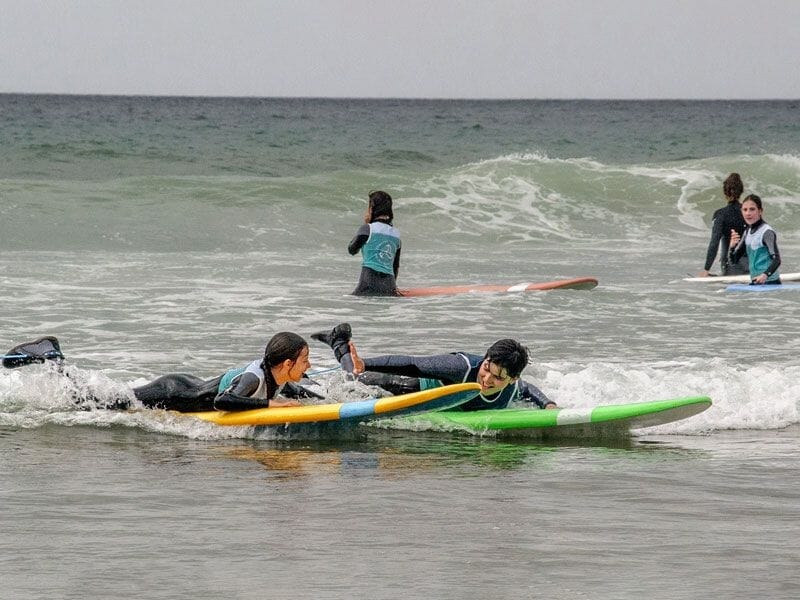 Deux enfants en combinaison de surf allongés sur leurs planches colorées dans l'eau, prêts à attraper une vague, avec d'autres surfeurs en arrière-plan.