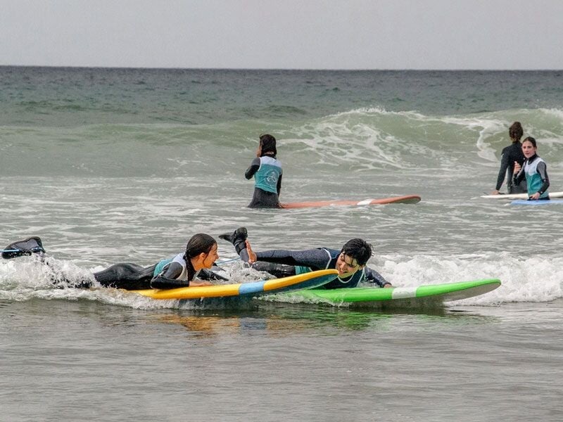 Deux enfants en combinaison de surf allongés sur leurs planches colorées dans l'eau, prêts à attraper une vague, avec d'autres surfeurs en arrière-plan.