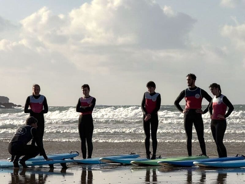Un groupe de six surfeurs en combinaison noire et rouge debout sur la plage avec leurs planches, écoutant un instructeur accroupi.