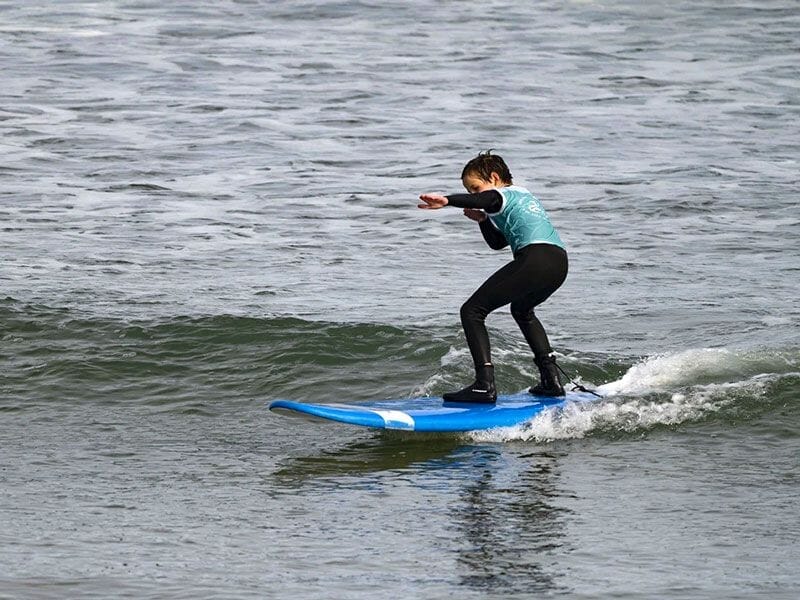 Un enfant en combinaison noire et bleu surfant debout sur une planche bleue, équilibré sur une petite vague dans l'océan.