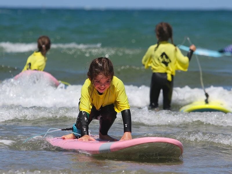 Une fillette souriante en combinaison jaune sur une planche rose dans l'eau peu profonde, avec deux autres enfants en arrière-plan.