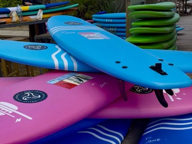 Planche de surf bleue et rose empilées sur un quai en bois avec plusieurs autres planches vertes en arrière-plan.
