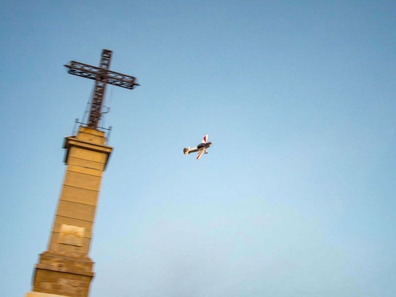 Petit avion de voltige volant près d'une grande croix sur un monument sous un ciel bleu clair.