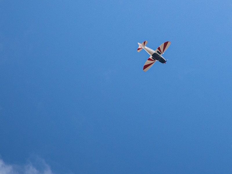 Avion de voltige vu de dessous en plein ciel bleu, ailes blanches avec motifs rouges.
