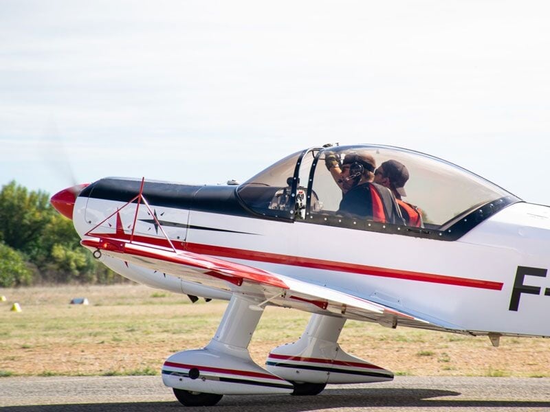 Avion de voltige blanc et rouge au sol avec deux personnes à bord, prêt au décollage.