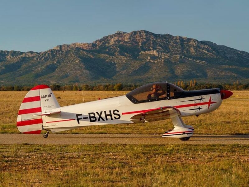 Avion de voltige blanc et rouge posé sur une piste avec une montagne en arrière-plan.