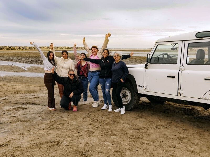 Un groupe de personnes souriantes pose joyeusement près d'un 4x4 blanc sur une plage sableuse au bord de l'eau.