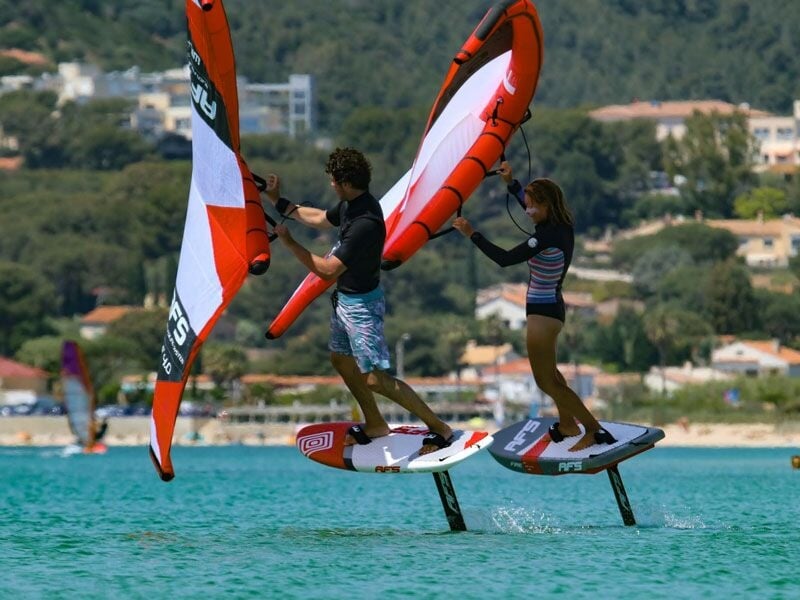Deux personnes pratiquent le wingfoil sur une eau turquoise avec des collines et des maisons en arrière-plan sous un ciel clair.