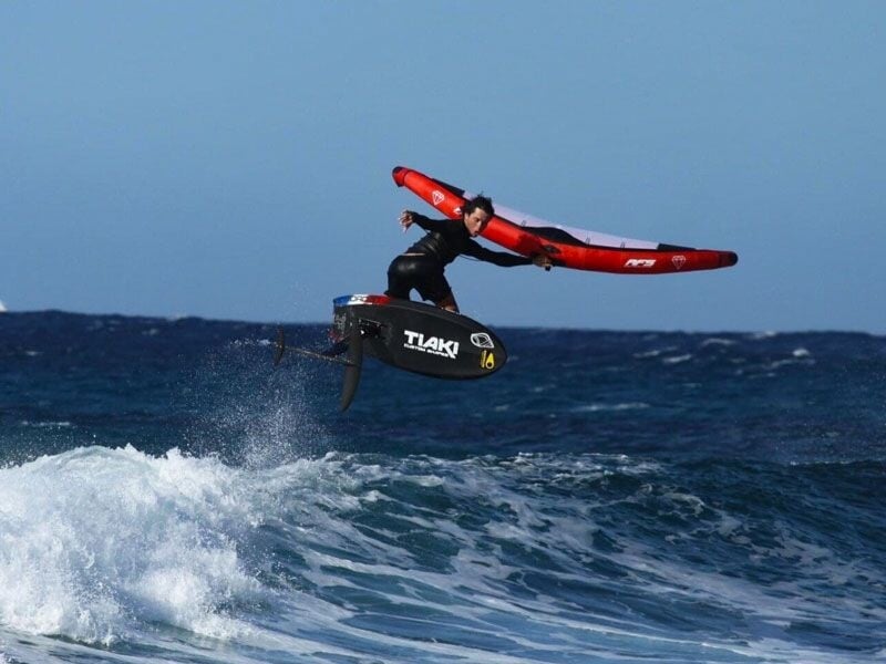 Un pratiquant de wingfoil effectue un saut spectaculaire au-dessus des vagues en mer agitée sous un ciel bleu clair.