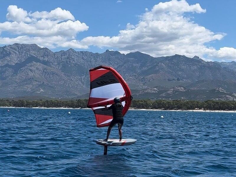 Un homme debout sur une planche de wingfoil navigue sur une mer calme avec des montagnes en arrière-plan sous un ciel nuageux.
