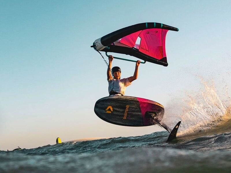 Un sportif en wingfoil saute au-dessus de l'eau au coucher du soleil, tenant une aile rouge et noire avec énergie.