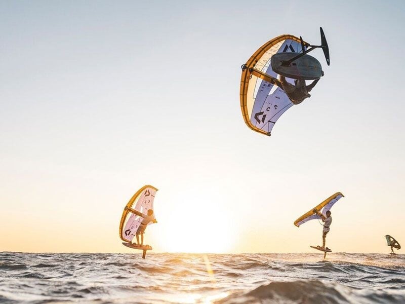 Plusieurs riders en wingfoil réalisent des figures aériennes au coucher du soleil sur une mer calme et dorée.
