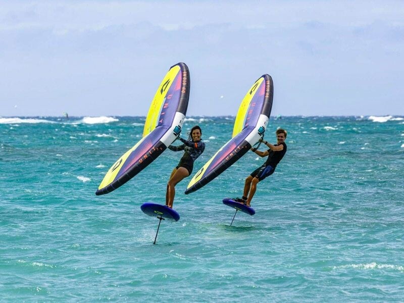 Deux personnes souriantes pratiquent le wingfoil côte à côte sur une mer turquoise avec des ailes jaunes et violettes.