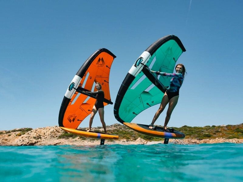 Deux femmes en wingfoil glissent sur l'eau turquoise près d'une côte rocheuse sous un ciel bleu clair et ensoleillé.