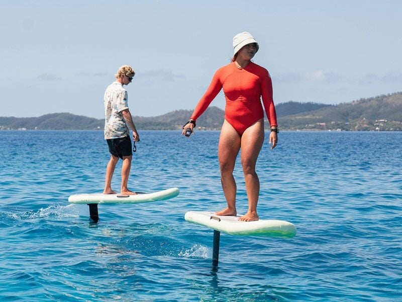 Deux personnes pratiquent le foil sur planche électrique sur une mer calme avec des collines en arrière-plan sous un ciel clair.